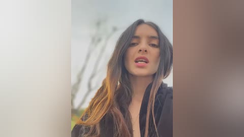 Media: A video featuring a young woman with long, wavy, dark hair, wearing a black jacket, with a blurred outdoor background.