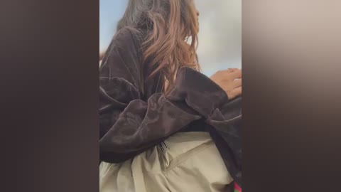 Media: Video of a person with long, wavy brown hair, wearing a black velvet jacket and beige pants, standing in a dimly lit room with blurred background.