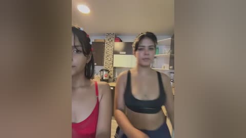 Media: Video of two young women in a home, one with short black hair, red top, and glasses, the other with dark hair, black sports bra, standing in a kitchen with cabinets and appliances visible.