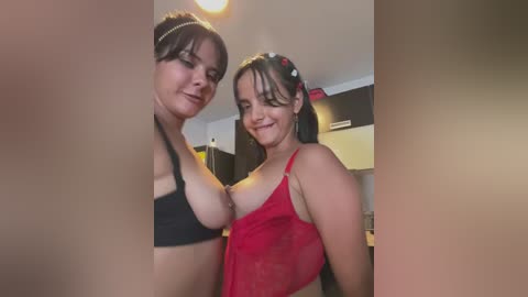 Media: Video of two women in lingerie, one in a black bra, the other in a red mesh top, smiling in a dimly lit room with kitchen appliances in the background.