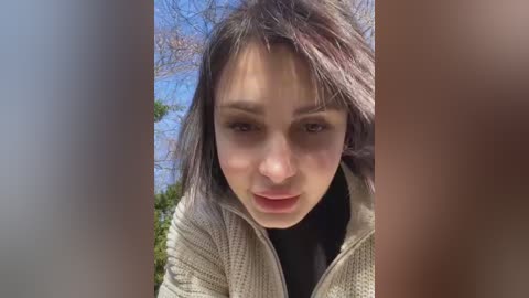 Media: Video of a young woman with light skin and dark hair, wearing a beige sweater, leaning forward with a concerned expression, outdoors with a blurred background.