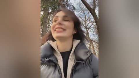 Media: Video of a smiling woman with long brown hair, wearing a grey puffer jacket, standing in a forest with bare trees and a cloudy sky.