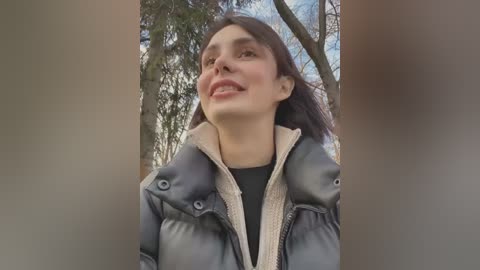 Media: Video of a smiling young woman with dark hair, wearing a black shirt and a dark puffy jacket, standing in a forest with tall trees and a blue sky.