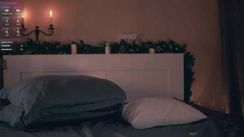 Video of a dimly-lit bedroom with a white headboard, a lit candle on a black wall sconce, and a bed with two pillows (one blue, one white). A garland of greenery and white lights decorates the headboard.