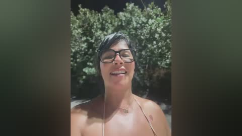 Video of a smiling, middle-aged woman with short, dark hair and glasses, wearing a pink bikini, set against a lush, green hedge background.