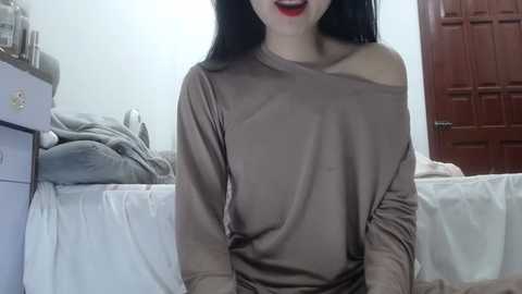 Media: Video of an Asian woman with long black hair, wearing a loose, off-the-shoulder brown top, sitting on a bed with white sheets, and a brown door in the background.