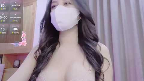 Media: Video of an East Asian woman with long black hair, wearing a white surgical mask, revealing cleavage in a light-colored top. Background features a wooden door and a digital display showing health metrics.