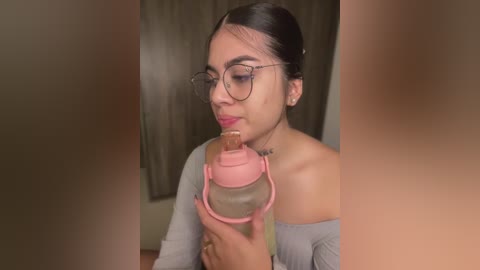 Media: A video of a young Latina woman with dark hair, glasses, and a grey off-shoulder top, drinking from a pink baby bottle in a dimly lit room.