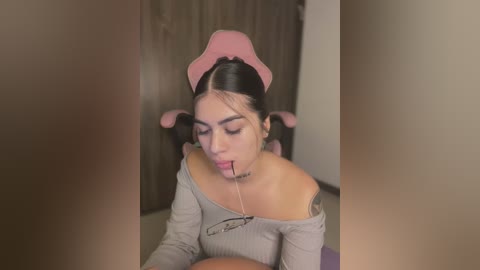 Media: Video of a woman with long black hair, wearing a pink headband and a grey off-shoulder top, sitting on a chair, with a pink lollipop in her mouth.