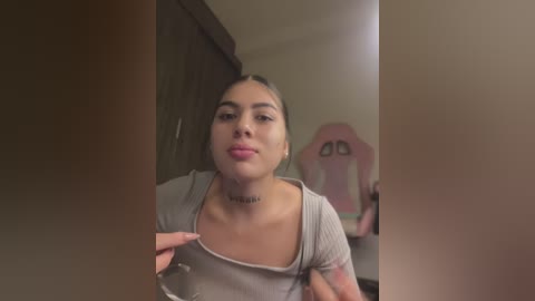 Media: Video of a young woman with light brown skin, dark hair pulled back, wearing a grey tank top. She has a tattoo on her neck and a pink gaming chair in the background.