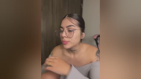 Media: Video of a young Latina woman with dark hair, wearing glasses, off-shoulder grey top, and tattoos. She's seated, looking down at her hands, in a dimly lit room with a wooden door in the background.