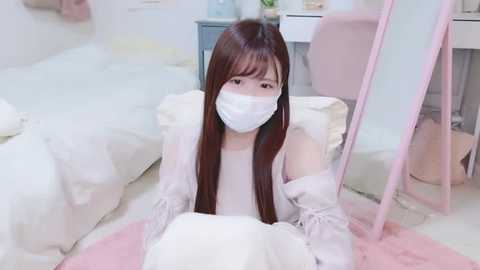 Media: Video of a young Asian woman with long brown hair and pale skin, wearing a white face mask and white robe, kneeling on a pink rug in a minimalistic, pastel-colored bedroom.