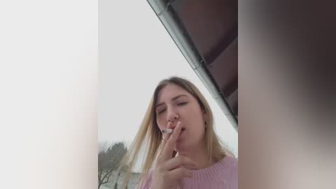 Media: Video of a blonde woman in a pink sweater, holding a cigarette, against a cloudy sky and a roof, her expression contemplative.