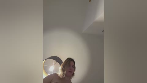 Media: A video of a naked woman with light skin and shoulder-length blonde hair, standing in a dimly lit room with white walls and a round, yellow lamp.