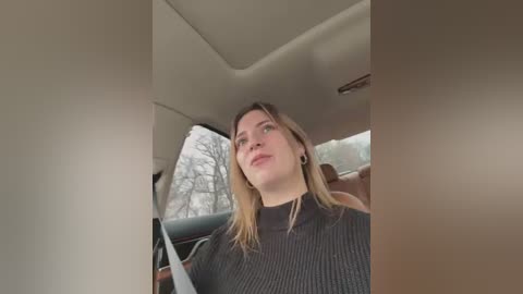 Media: Video of a young Caucasian woman with shoulder-length blonde hair, wearing a black ribbed turtleneck sweater, sitting in a car with a snow-covered tree outside the window.