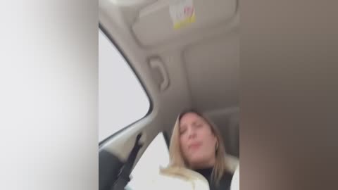 Media: Video of a woman with long blonde hair, wearing a white jacket, asleep in a car seat. The car interior is beige with a sunroof, and the background is blurred.