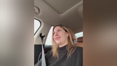 Media: Video of a smiling woman with long, light brown hair, wearing a dark gray knitted sweater, sitting in a modern car with beige interior, looking out the window.