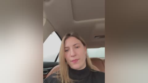 Media: Video of a smiling young woman with light skin and long blonde hair, wearing a black turtleneck, seated in a car with beige interior.