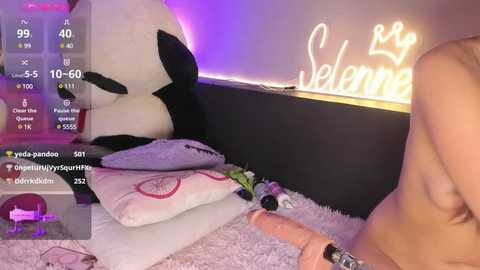 Media: Video of a topless woman on a bed with a stuffed panda, purple lighting, and \"Selena\" neon sign in background.