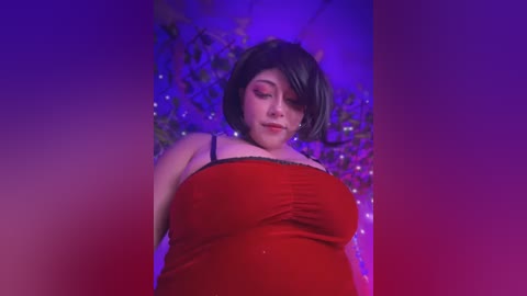 Media: Video of a plus-size woman with short dark hair, wearing a red spaghetti-strap dress, lying on a bed with purple and blue lighting, creating a dramatic, sensual atmosphere.