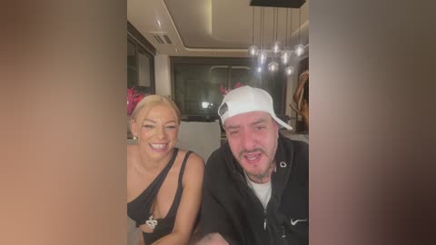 Media: Video of a smiling blonde woman in a black dress with a red flower, and a bearded man in a white cap and black jacket, indoors with modern lighting.