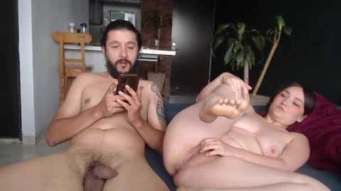 Media: Video of a bearded, nude man with a circumcised penis, holding a smartphone, and a nude, fair-skinned woman with medium breasts, lying on a bed, both indoors.