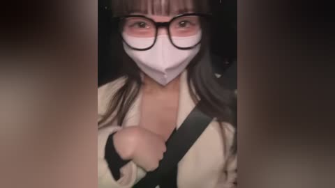 Media: Video of a woman with large, round glasses, white face mask, and brown hair, wearing a beige coat with a black shoulder strap, driving in a car.