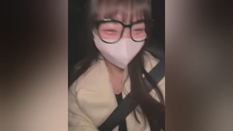 Media: Video of an Asian woman wearing glasses, a white mask, and a beige jacket, with a seatbelt across her chest, sitting in a dark car.