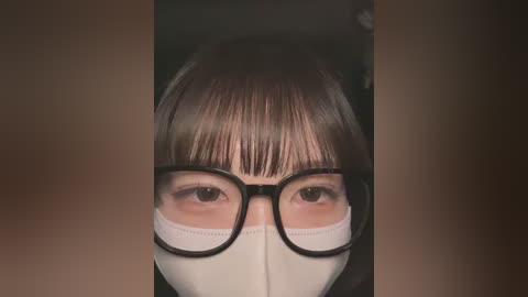 Media: Video of an Asian woman with straight, dark hair and bangs, wearing black-framed glasses and a white face mask, with a blurred, dark background.