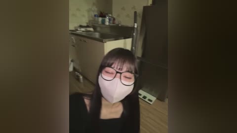 Media: A video shows a woman with long dark hair, wearing glasses and a white mask, standing in a dimly lit kitchen with a sink, stove, and refrigerator.