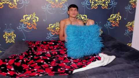 Media: Video of a shirtless, fit, young man with short brown hair, seated on a bed with a red and black patterned blanket. He holds a blue, fluffy pillow, with a dark wallpaper featuring yellow and blue floral designs in the background.