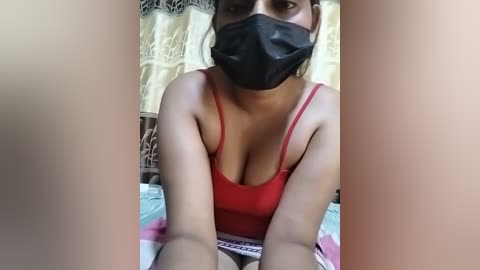 Media: Video of a woman with light brown skin, wearing a black face mask and red tank top, lying on a bed with patterned sheets.