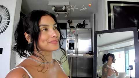 Media: Video of a smiling woman with dark hair, wearing a white tank top, standing in a modern kitchen with a stainless steel refrigerator, mirrored wall, and \"Silvia Luna\" logo.