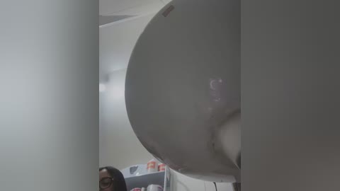 Media: A video showing a large, white, round object, likely a satellite dish, partially visible in a room with a woman wearing glasses and a black top, standing near a white wall and a door.