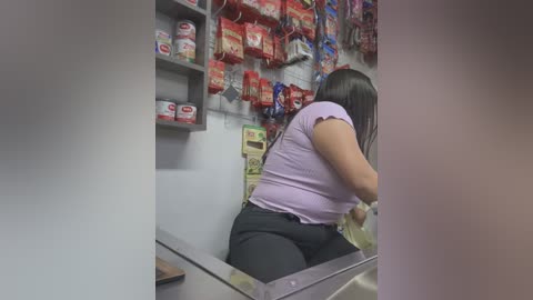 Media: A video of a Latina woman with medium skin tone, long black hair, and a purple top, scanning groceries in a dimly lit store. Red packages of chips hang on the shelves behind her.