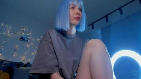 Media: Video of a fair-skinned woman with short blue hair, wearing a loose grey t-shirt, sitting in a dimly lit room with string lights and a glowing ring light.