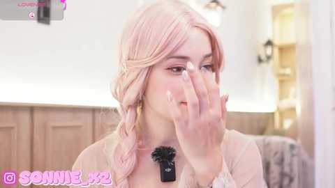 Media: Video of a fair-skinned woman with pink hair, wearing a pastel top, applying makeup with a black brush, in a cozy, softly lit room.