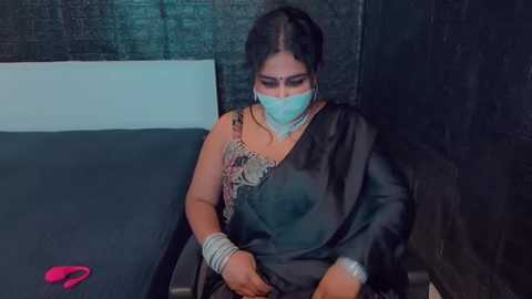 Media: Video of a woman in a black sari with a gold border, wearing a face mask, silver bangles, and earrings, sitting on a bed with a dark headboard, in a dimly lit room with a brick wall.