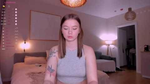 Media: Video of a young woman with straight brown hair, wearing a gray tank top, sitting on a bed in a modern, dimly lit bedroom.