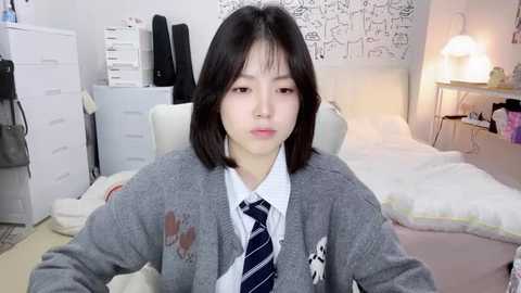 Media: Video of an East Asian woman with straight black hair, wearing a gray cardigan over a white shirt and navy striped tie, sitting in a modern, minimalist bedroom with white furniture, black-and-white patterned wallpaper, and a white bed.