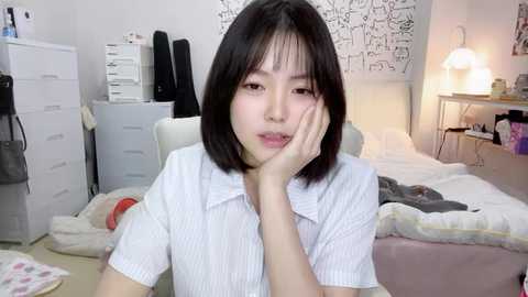 Media: Video of a young Asian woman with straight black hair, resting her chin on her hand, wearing a white striped polo shirt, in a cluttered, softly lit bedroom with white furniture, a bed, and wall art.