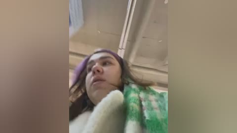 Media: Video of a young woman with light skin and brown hair, wearing a purple hat and green plaid jacket, taken from a low angle, capturing a casual, indoor setting.