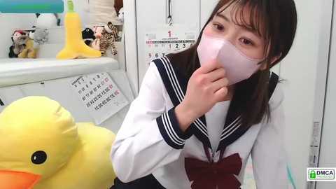 Video of a young Asian woman in a school uniform, wearing a surgical mask, holding a stuffed yellow duck, in a brightly lit classroom with white walls and a calendar.