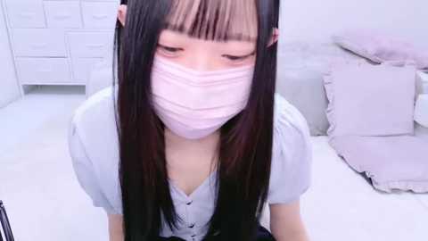 Media: Video of an East Asian woman with long black hair, wearing a light blue blouse and a pink face mask, kneeling in a minimalist, white-walled bedroom with white drawers and a bed.