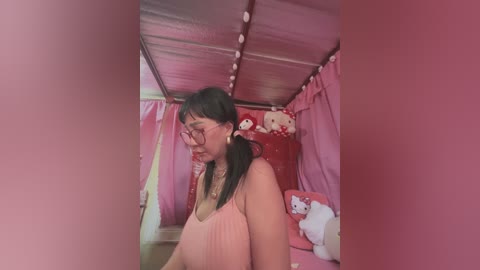 Media: Video of a young woman with glasses, pink top, and pigtails in a pink-themed room with plush toys and string lights.