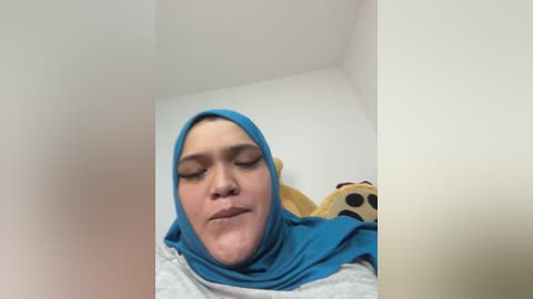 Video of a person with fair skin and blue hijab, eyes closed, wearing a light gray shirt, lying on a white surface, with a stuffed bear nearby.