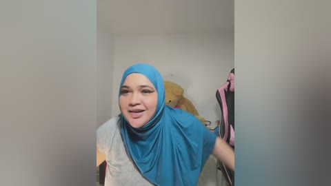 Video of a light-skinned woman with a blue hijab, wearing a gray shirt, smiling, standing in a cluttered room with beige walls and hanging clothes.