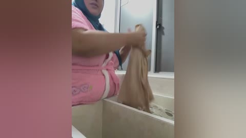 A video shows a person in a pink robe with a blue hijab, washing a brown cat in a beige bathtub, in a modern bathroom with a glass shower enclosure.