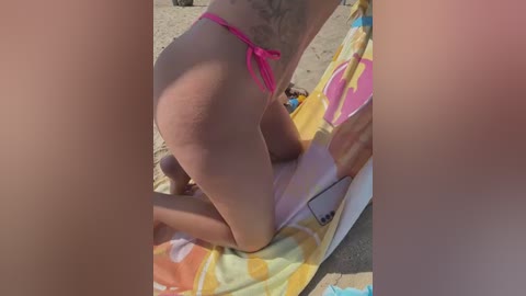 Media: A video of a topless woman with light skin, wearing a pink thong bikini, kneeling on a colorful towel on a sandy beach.