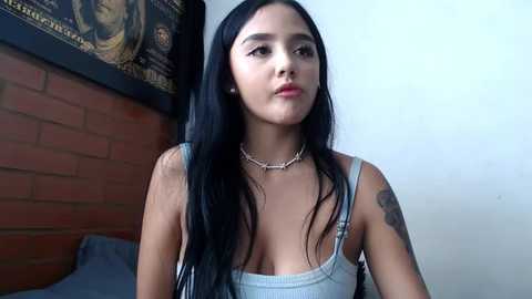 Media: Video of a Latina woman with long black hair, medium skin tone, medium build, wearing a light grey tank top, sitting indoors near a brick wall.